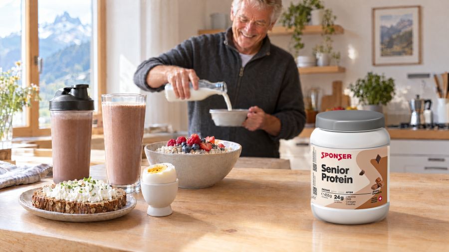 Bright kitchen with an older man in the background; a chocolate shake, porridge, an egg, bread with cottage cheese and Sponser Senior Protein sit on the table.