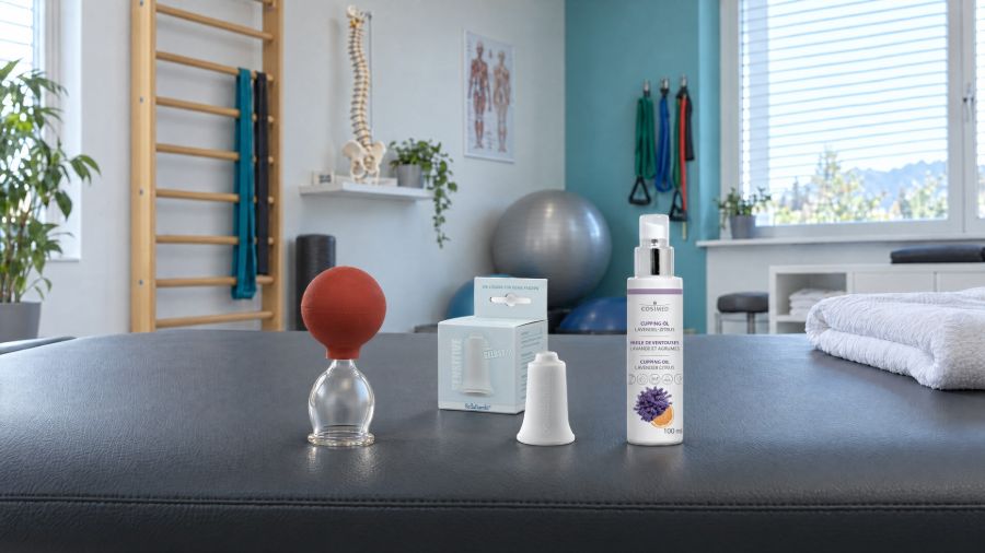 Lauschaer Schröpfglas mit rotem Saugball, weisser BellaBambi-Cup und cosiMed Cupping-Öl auf einer Behandlungsliege in einer Physiopraxis.