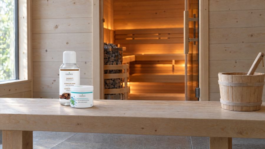 Holzbank im Vorraum: links cosiMed Saunaduft Zedernholz 200 ml und davor eine Mentholdose 50 g, rechts Holzkübel mit Kelle. Durch die offene Tür ist der Saunaofen zu sehen.