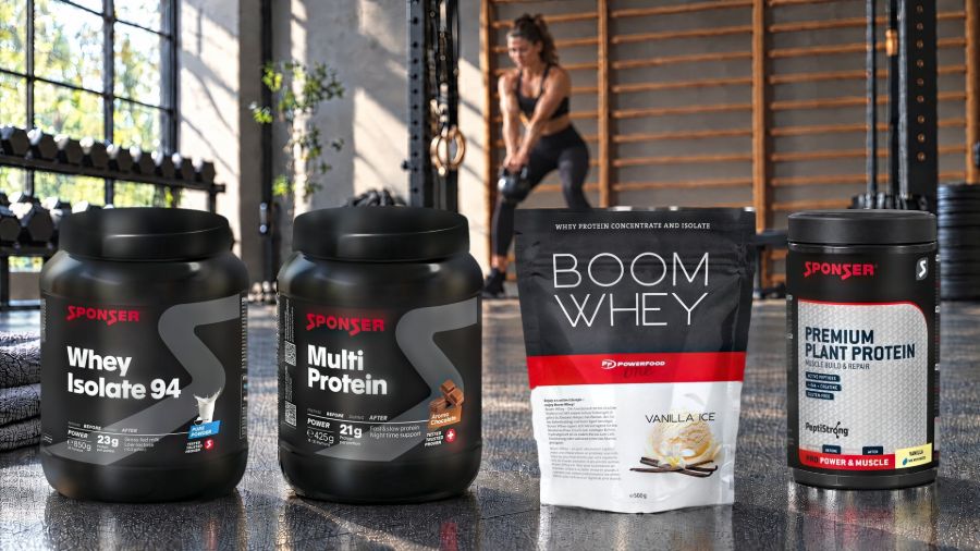 Four protein powders on the floor of a gym with dumbbells and wall bars, a woman training with a kettlebell in the background: Sponser Whey Isolate 94, Sponser Multi Protein, Powerfood One Boom Whey and Sponser Premium Plant Protein.
